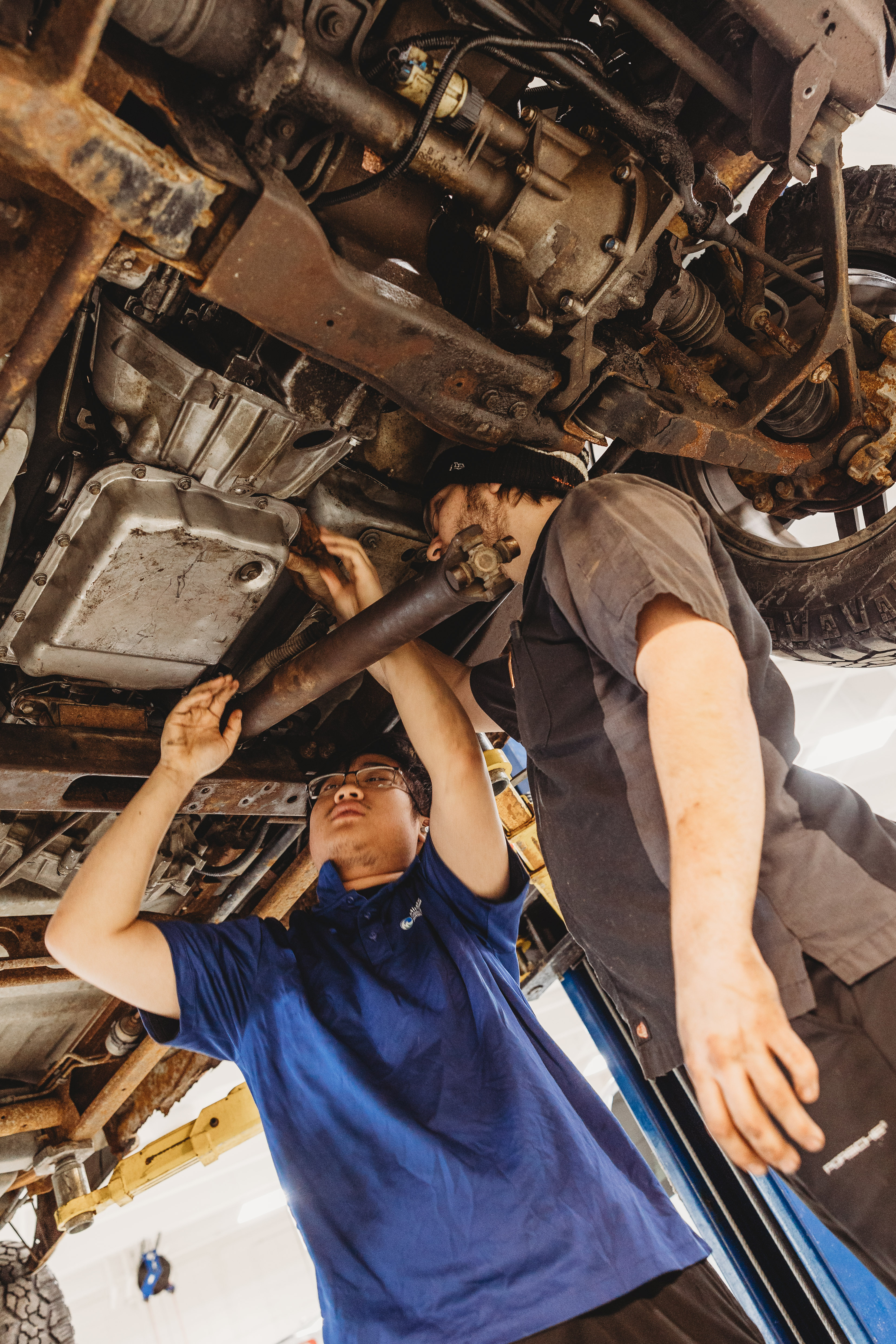 Two M State automotive students in blue M State shirts stand underneath a lifted car in the transportation lab on the Moorhead campus, using hand tools to make repairs to the underside of the vehicle.