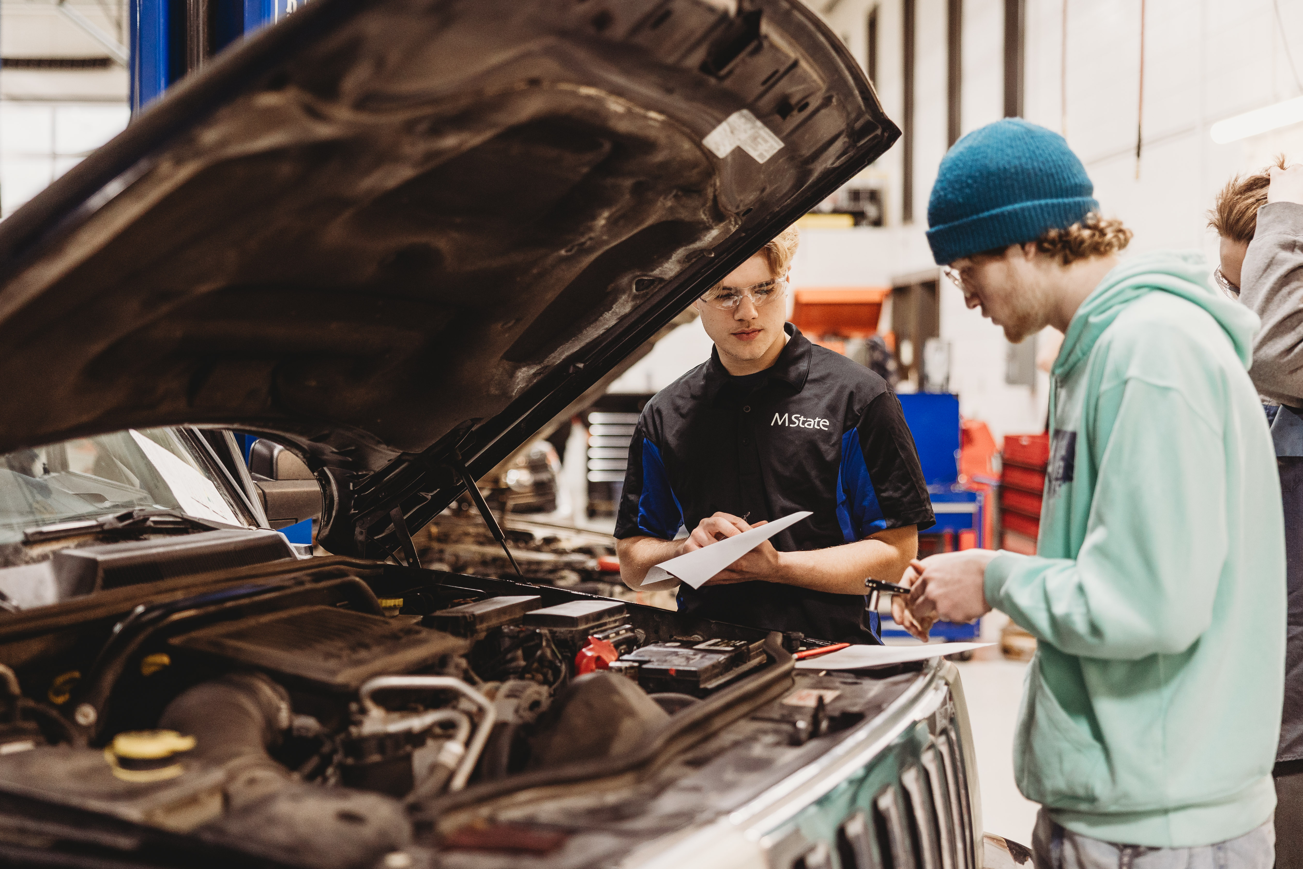 Two students in M State's automotive program stand in front of a vehicle with the hood up, examining the engine to diagnose problems.