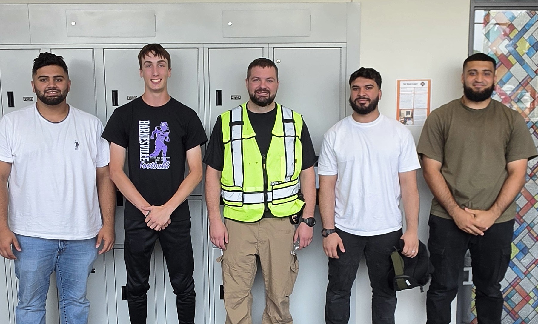 Four M State Criminal Justice students pose with a police officer wearing a reflective yellow vest in a high school hallway with gray lockers in the background during an active shooter training in Moorhead.