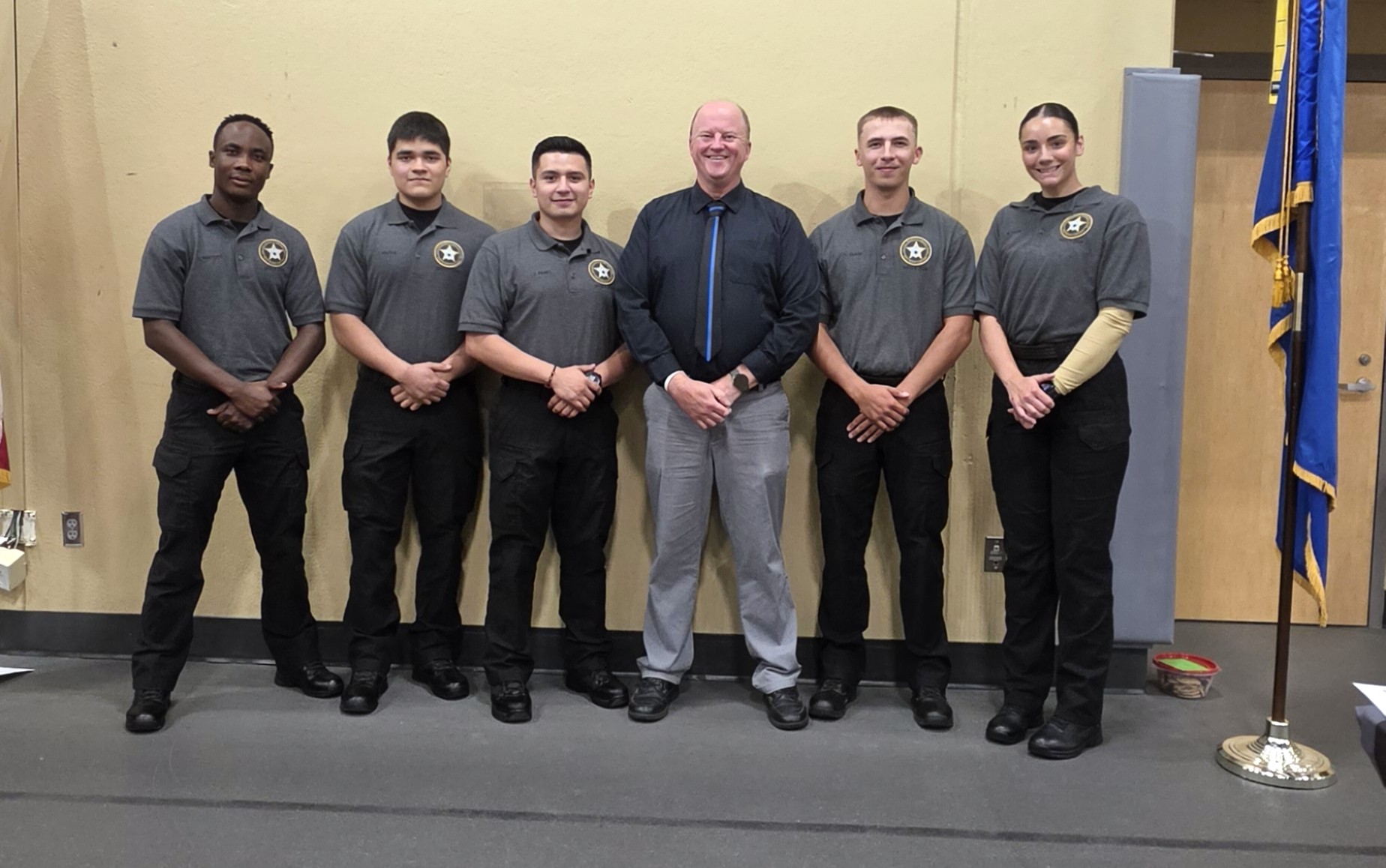Five M State Criminal Justice students, wearing matching black pants and gray shirts with star badges on the chest, post with instructor Jeff Nelson after completing a skills training.