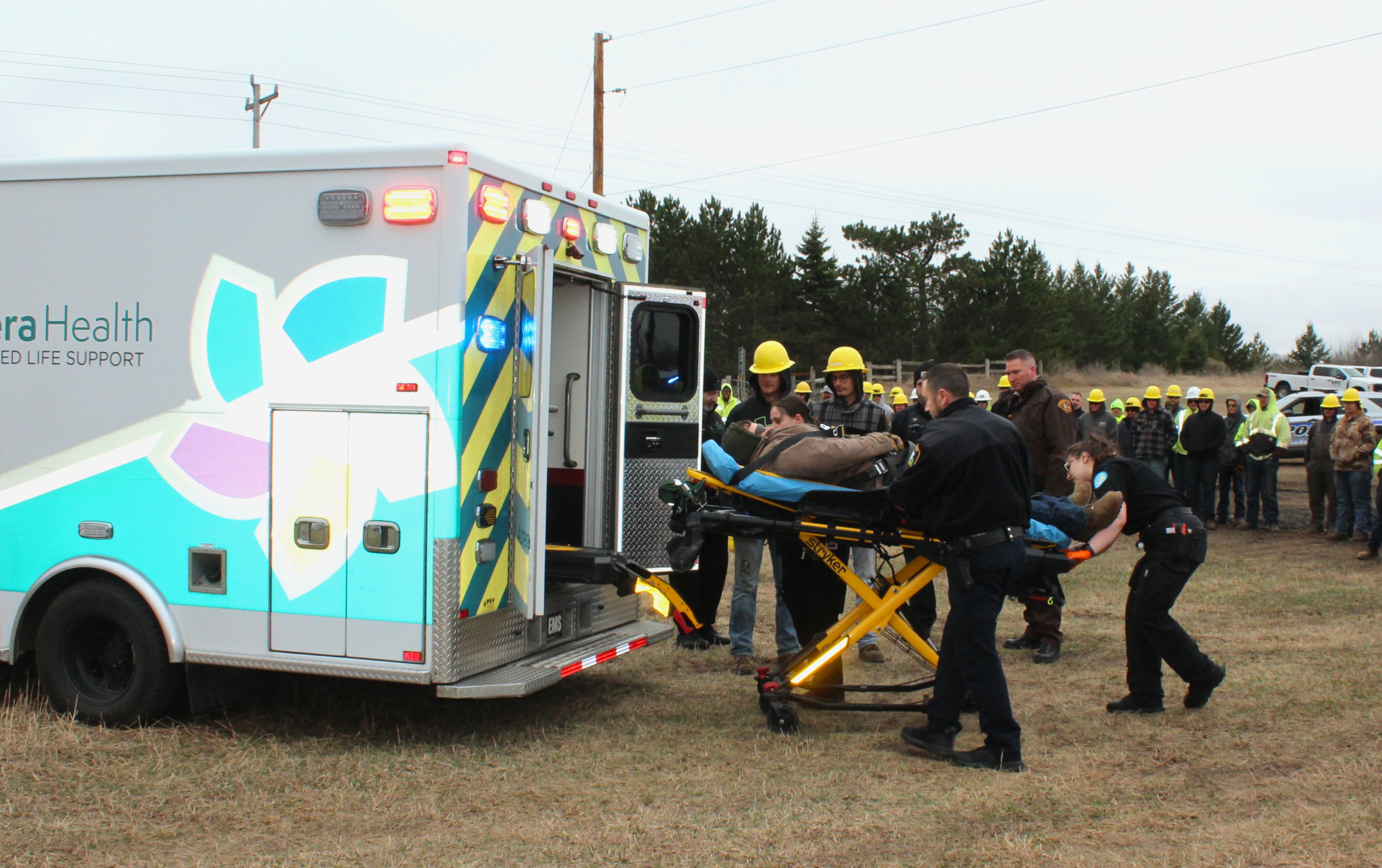 Several emergency responders, offers and M State line worker students lift an "injured patient" on a stretcher into an ambulance as part of an emergency training drill at the M State line worker training field on Friday, April 17, as a group of students and other participants look on.