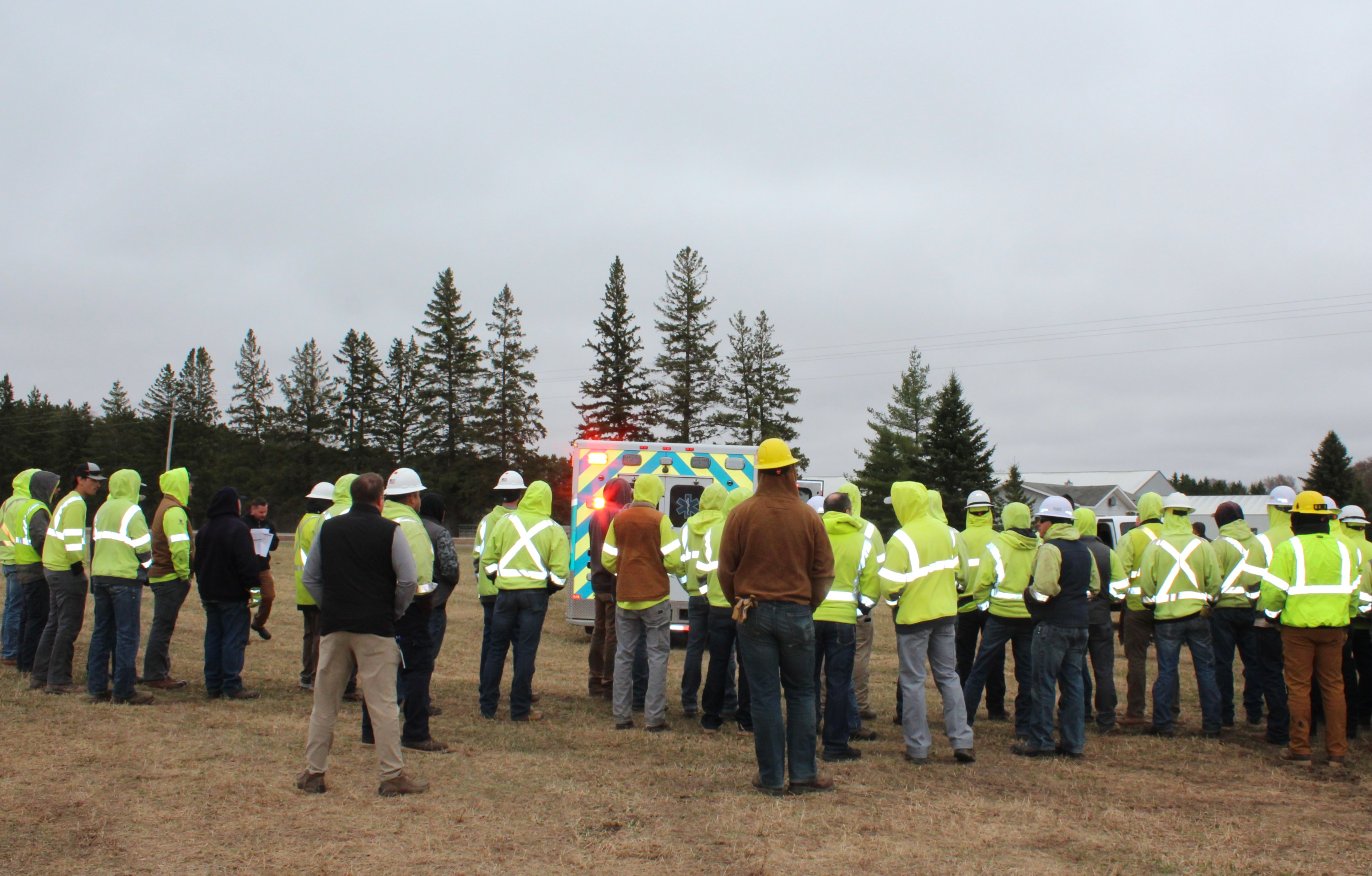 A group of about 30 line worker students wearing yellow reflective jackets and hard hats, along with several other participants in a large-scale emergency drill training exercise, look on as an ambulance transports an "injured worker" from the scene.