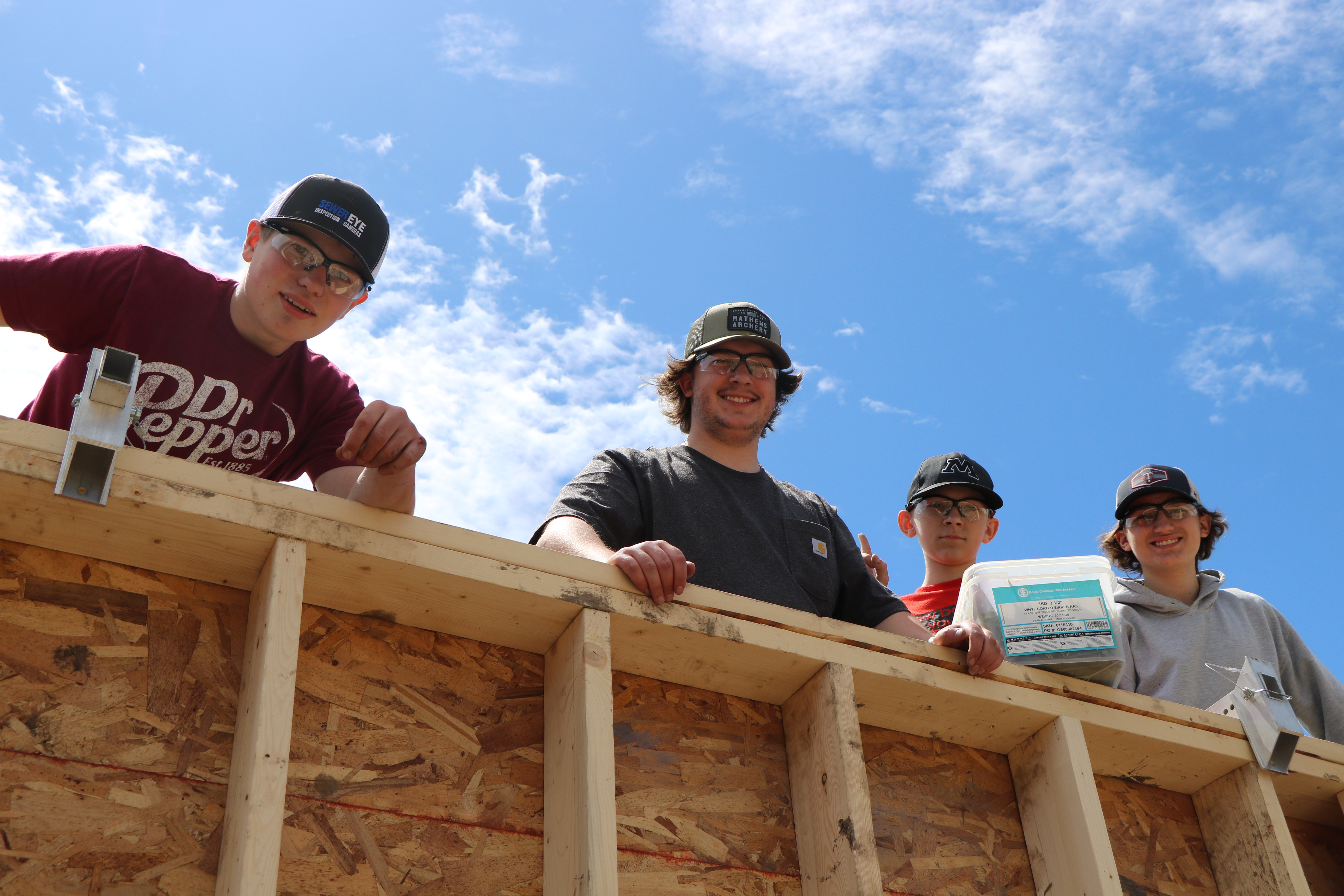 Four male youth, standing together at the 2024 Herdina Construction Trades Camp, look down from behind an under-construction wall and smile at the camera.