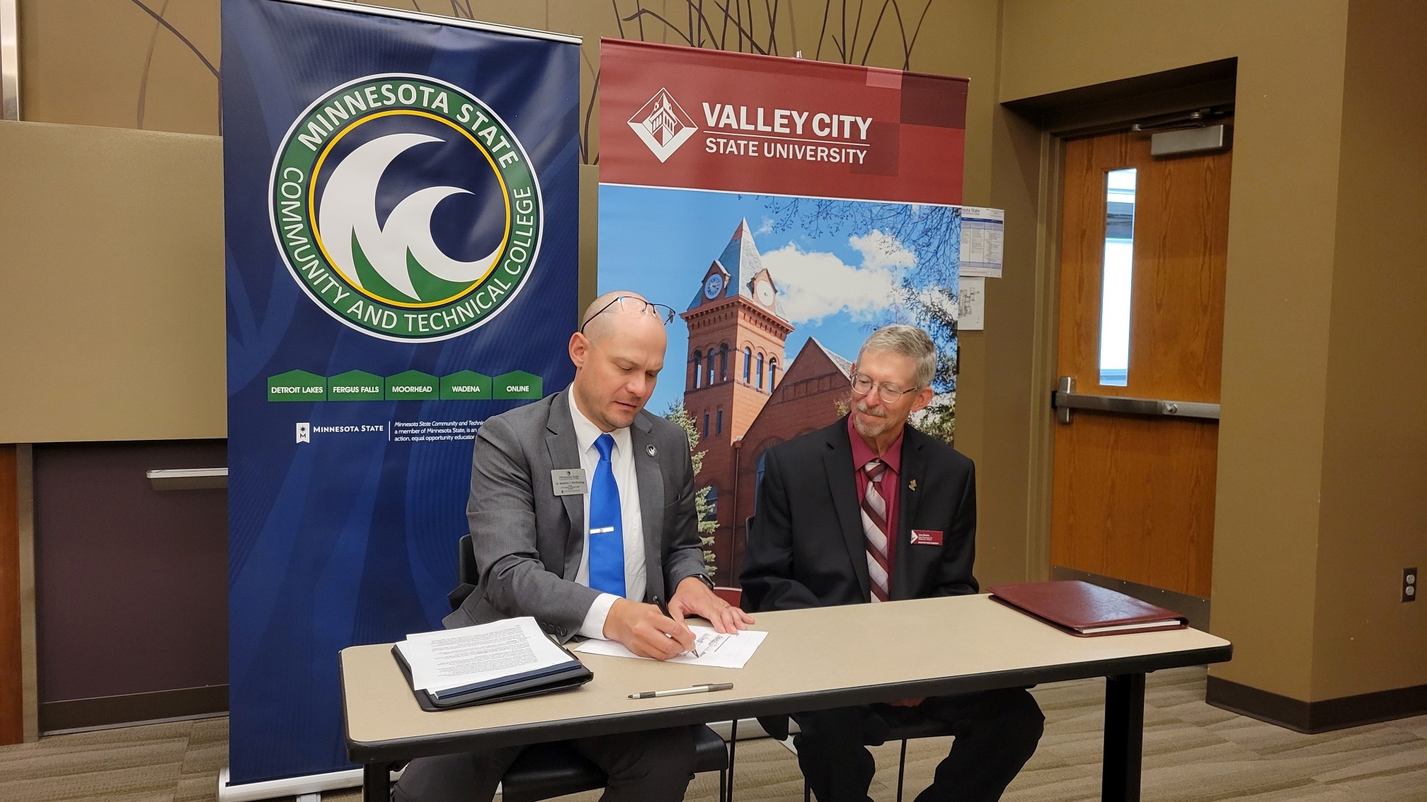 Two college leaders from M State and VCSU sit at a table reviewing an agreement between the schools.