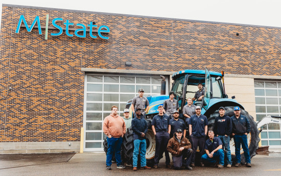 Photo of diesel students gathered around a tractor with the M State building in the background