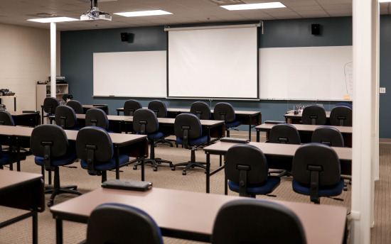 A traditional-style classroom with several tables and chairs with three projector screens in the front of the room