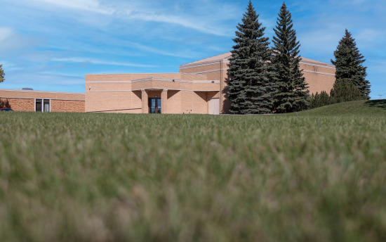 Exterior photo of M State-Detroit Lakes featuring a large grassy field and mature pine trees