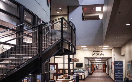 Interior photo showcasing the entrance to the library which is surrounded by windows, a metal staircase and a long hallway with a sign over the doorway that welcomes you to M State