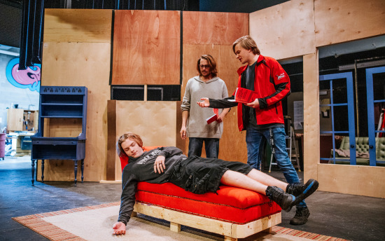 three people on stage in a room with a red couch