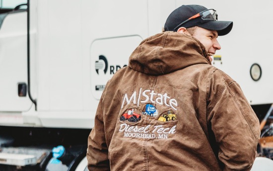 Student wearing an M State diesel tech jacket smiles as he walks toward a semi truck