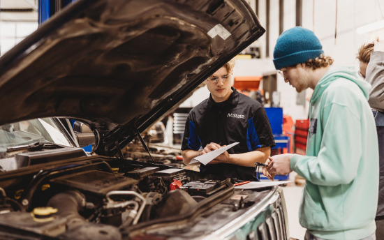 Two automotive students peer under the hood of a vehicle to examine the engine in class.
