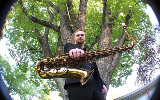 M State music graduate Andrew Danielson poses with his saxophone, looking down at the camera and holding his sax out in front of him with a large tree behind him