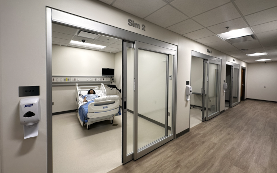 A scene inside the Health Sciences Simulation Center at M State's Moorhead campus, looking down a hallway with three simulation rooms with sliding glass doors, white walls, and hospital beds and equipment inside the rooms.