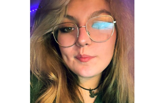 Head shot of a woman with blond hair and silver-rimmed glasses, with a closed-mouth smile and choker-style necklace