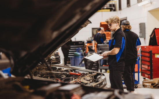 An M State automotive student, in a blue and black M State auto shirt, looks under the hood of a car in the Transportation Center at the Moorhead campus.