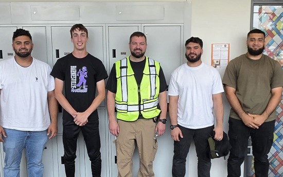 Four M State Criminal Justice students pose with a police officer wearing a reflective yellow vest in a high school hallway with gray lockers in the background during an active shooter training in Moorhead.