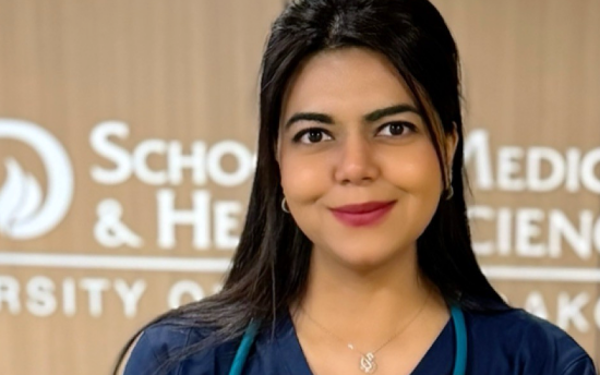 Head shot of an M State nursing graduate with long dark hair and dark blue scrubs, with a hospital hallway sign in the background