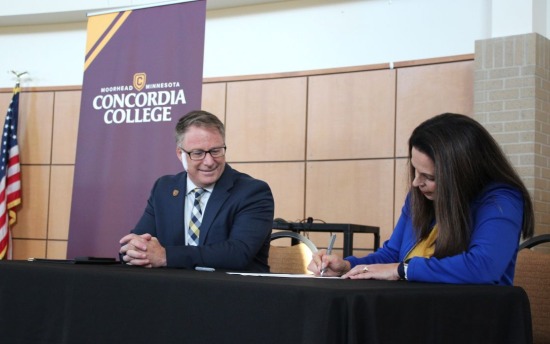Concordia College President Colin Irvine and M State President Carrie Brimhall sit at a table on the M State Fergus Falls campus, signing a new partnership agreement and smiling.