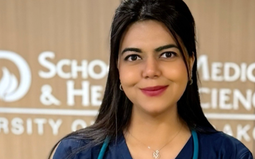 Head shot of an M State nursing graduate with long dark hair and dark blue scrubs, with a hospital hallway sign in the background