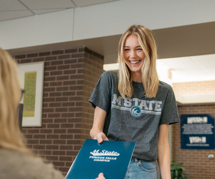 A person in an M State T-shirt accepts a folder from an M State employee