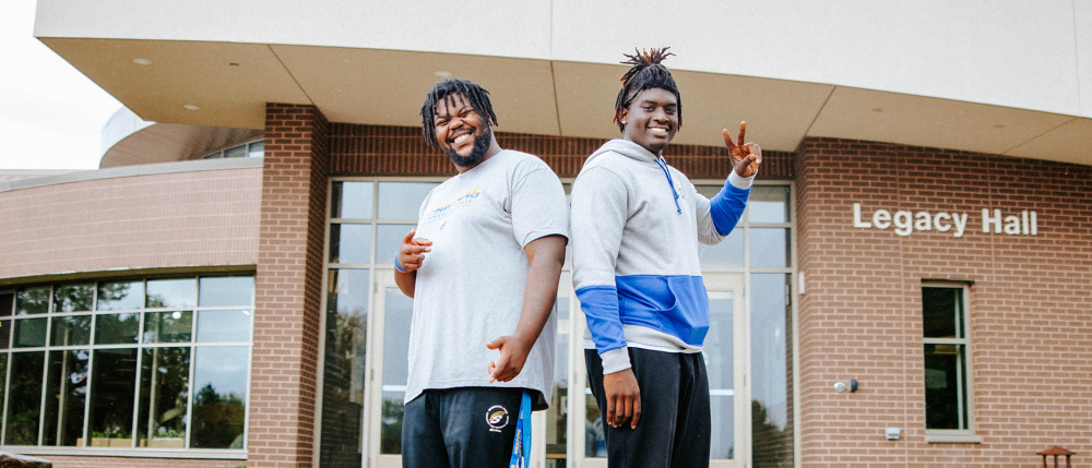 Two Men Smiling in front of brick round Legacy Hall on Fergus Fall campus