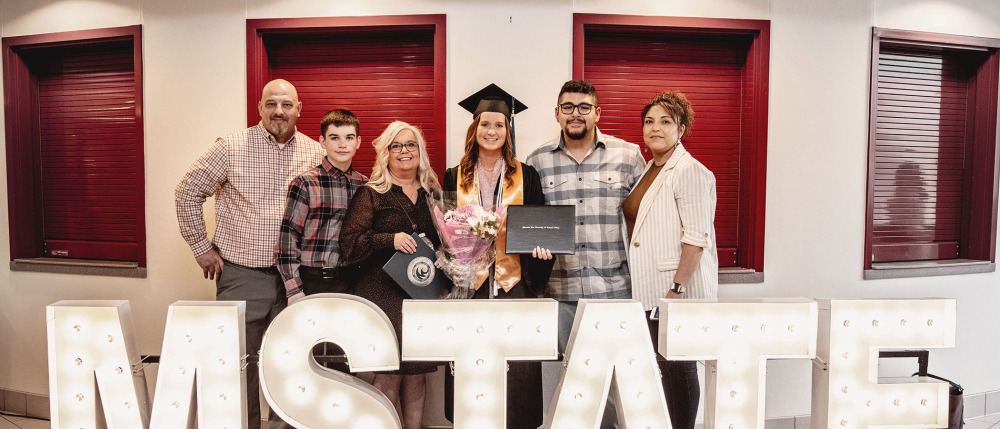 An M State student and their family stand behind an M State sign