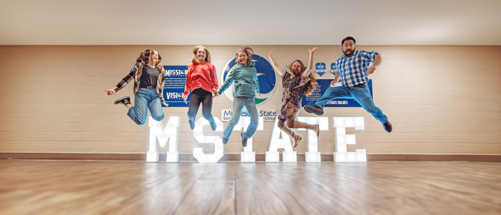 Four college students jump above an M State lighted sign. 