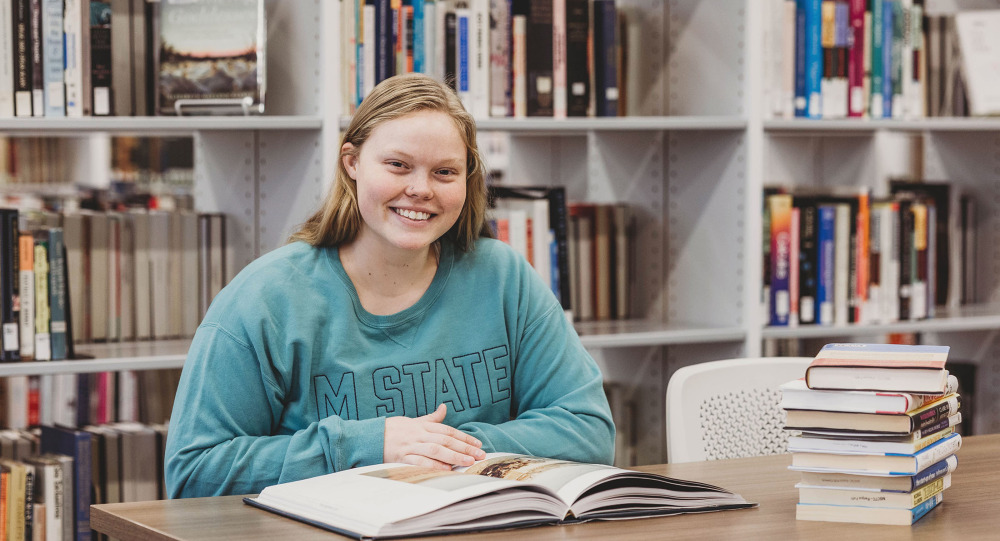 A student in a blue M State sweatshirt in the library smiles